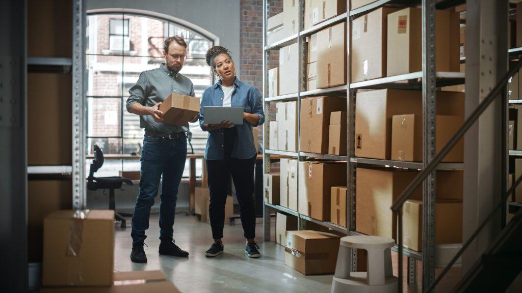 Two people stand in a warehouse surrounded by shelves filled with cardboard boxes; one holds a box, and the other holds a tablet, as they appear to discuss inventory or logistics in the brightly lit room.
