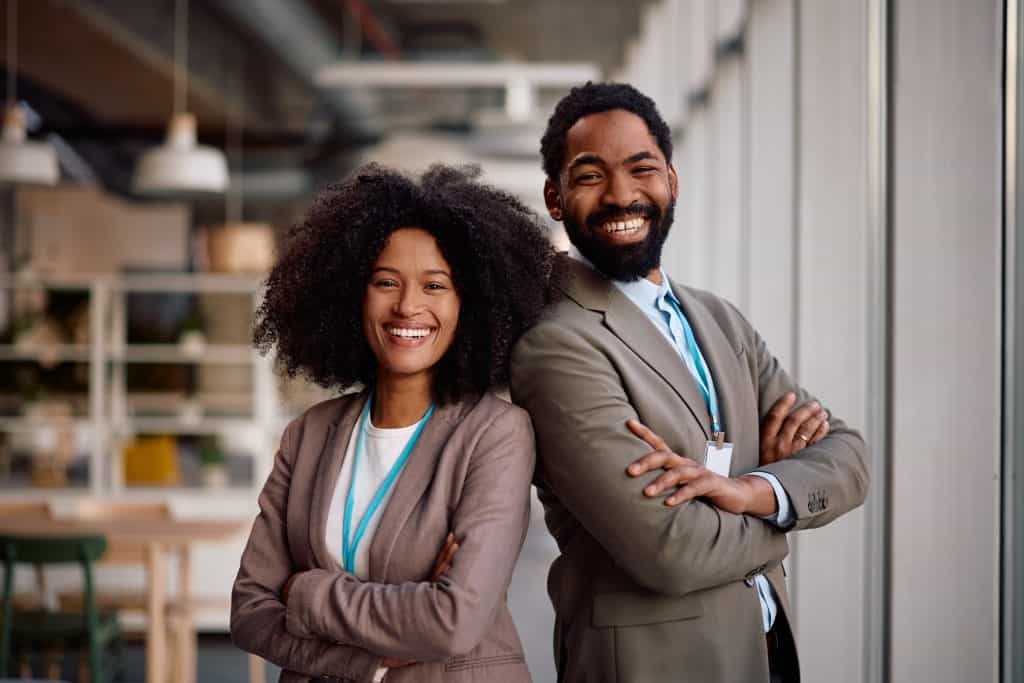 Two professionally dressed people, a woman and a man, stand side by side with arms crossed, smiling confidently in a modern office space with large windows.