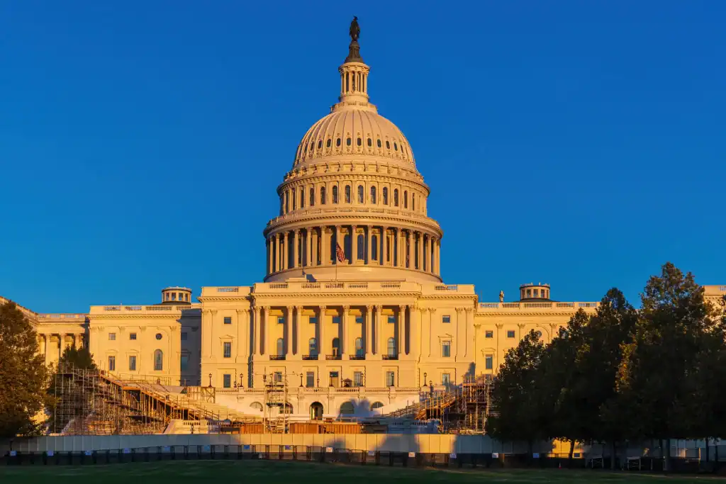 The United States Capitol building in Washington, D.C., illuminated by golden sunlight, with scaffolding around the lower steps and trees on both sides under a clear blue sky.