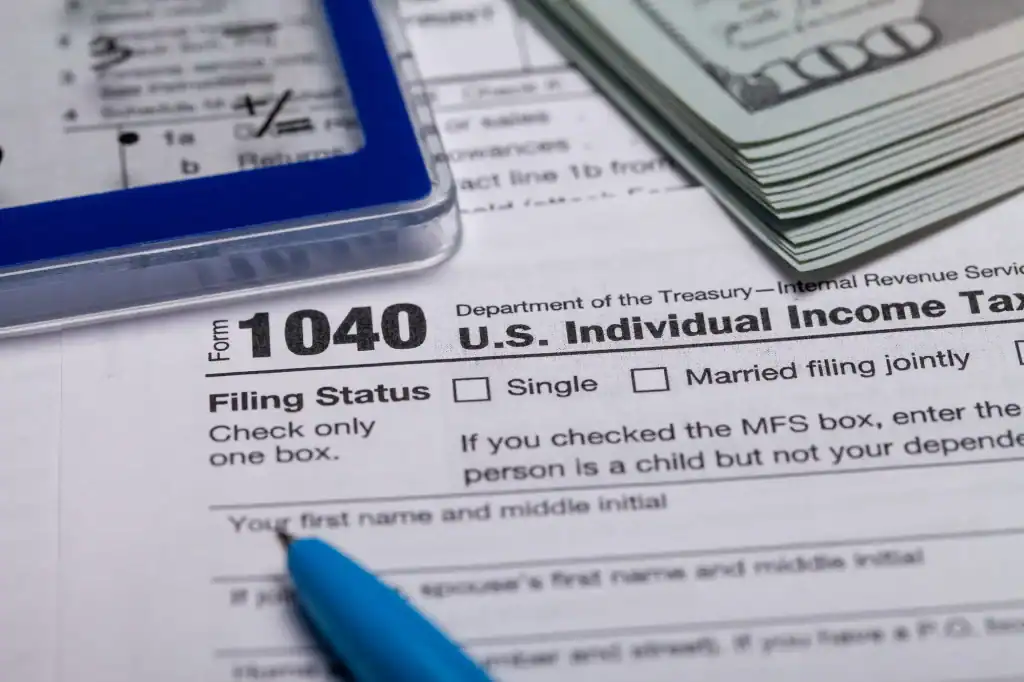 Close-up of a U.S. 1040 income tax form with a blue pen, a pile of hundred-dollar bills, and a calculator partially visible on the side. The tax form’s filing status section is in focus.