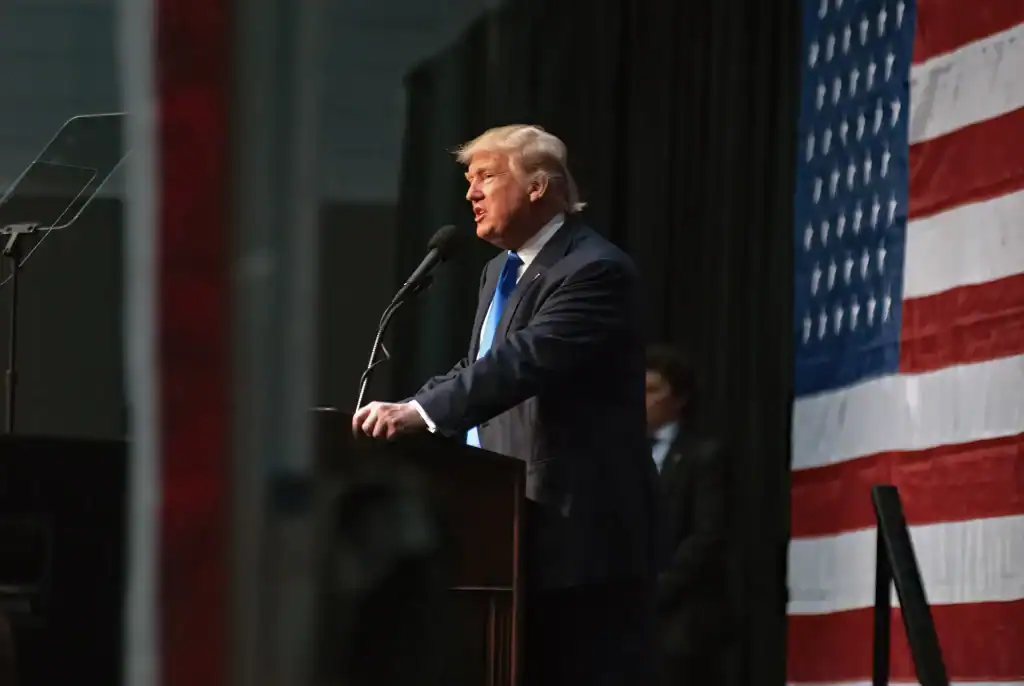 A man in a suit speaks at a podium with a microphone, standing in front of a large American flag.