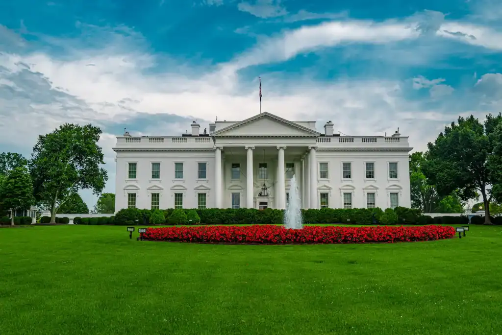 The White House, a large white neoclassical building, stands behind a green lawn and a circular flower bed with red flowers and a fountain, under a partly cloudy blue sky.